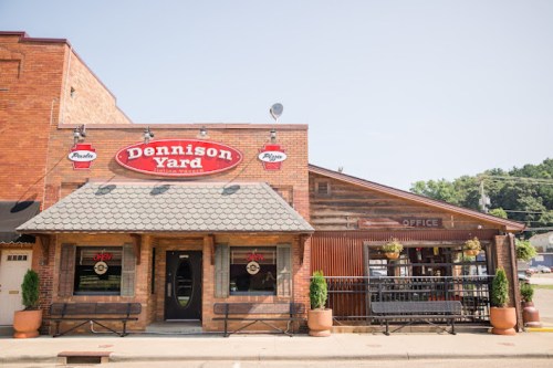 Exterior of Dennison Yard restaurant, featuring a brick facade, a sign, and outdoor seating on a sunny day.