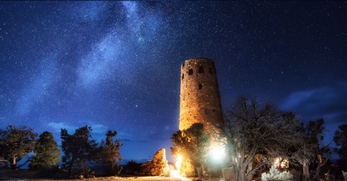 A stone tower illuminated at night under a starry sky, surrounded by trees and rocky terrain.