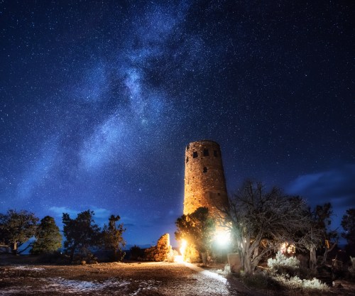 A stone tower illuminated at night under a starry sky, with trees surrounding it and a hint of the Milky Way visible.