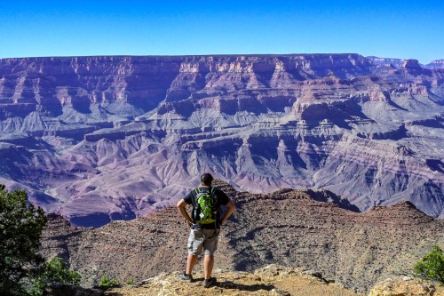 A hiker stands on a rocky ledge, gazing at the vast, colorful layers of the Grand Canyon under a clear blue sky.