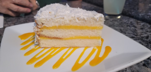 A slice of coconut cake with layers of yellow filling, served on a plate with a drizzle of mango sauce.