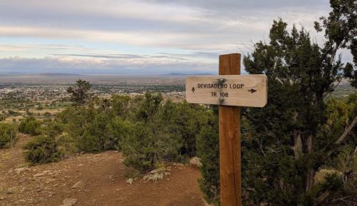 Wooden signpost for Devisadero Loop Trail 108, overlooking a valley with trees and distant mountains under a cloudy sky.
