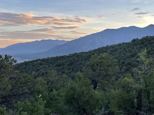 A scenic view of mountains at sunset, with lush green trees in the foreground and colorful clouds in the sky.