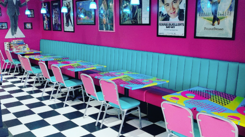 Colorful diner interior with pink walls, teal seating, and patterned tables, featuring black and white checkered flooring.
