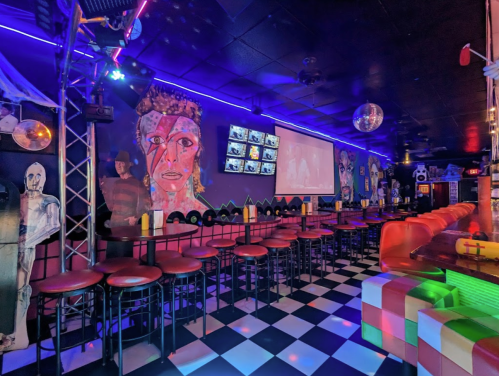 A vibrant bar interior with colorful murals, checkered floor, and illuminated seating under blue and purple lights.