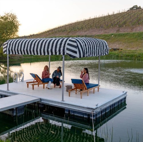 Three women relax on a striped canopy dock by a serene lake, surrounded by vineyards at sunset.