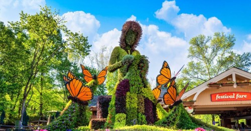 A large topiary figure surrounded by colorful flowers and butterflies, set against a blue sky and green trees.
