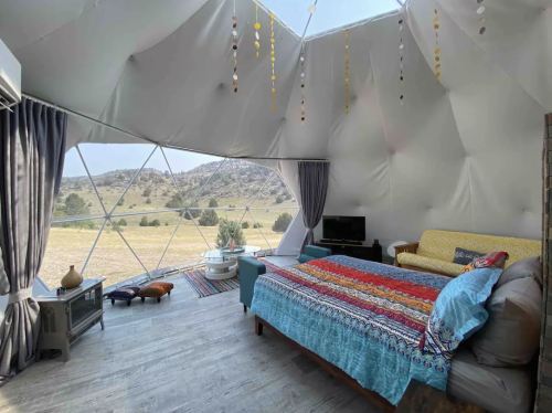 Interior of a geodesic dome with a colorful bedspread, sofa, and large windows showcasing a scenic landscape.