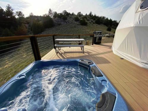 A hot tub on a wooden deck with a grill and picnic table, surrounded by greenery and hills under a cloudy sky.