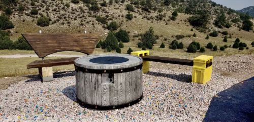 A circular wooden structure surrounded by colorful gravel, with two benches nearby, set against a hilly landscape.