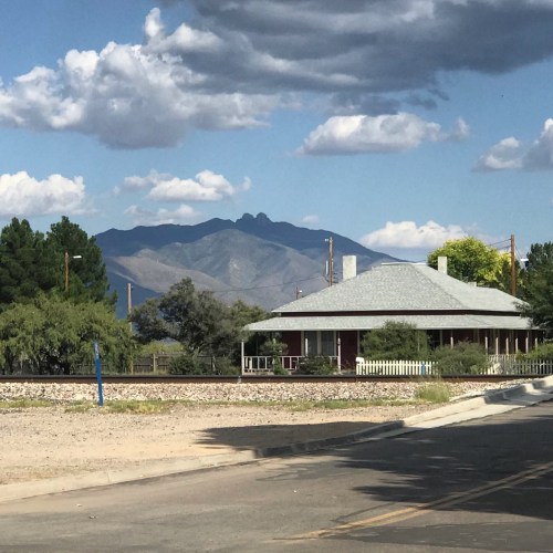 A quaint house with a gray roof sits near a road, with mountains and a cloudy sky in the background.