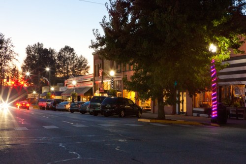A quiet street at dusk, lined with shops and trees, illuminated by streetlights and colorful decorations.