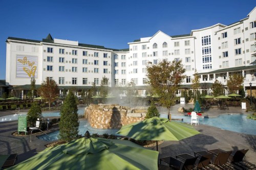 A resort with a large pool, hot tubs, and green umbrellas, surrounded by trees and a modern building under a clear blue sky.