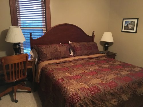 Cozy bedroom with a large bed, decorative quilt, two lamps, a wooden chair, and a framed picture on the wall.