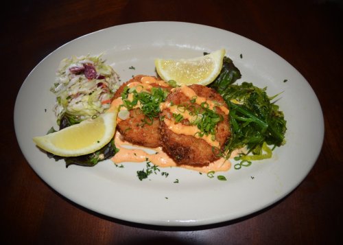 A plate featuring two fried crab cakes with sauce, lemon wedges, and a side of coleslaw and greens.