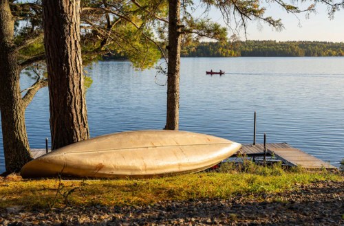 A canoe rests on the shore by a calm lake, with a dock and a person in a kayak in the background.