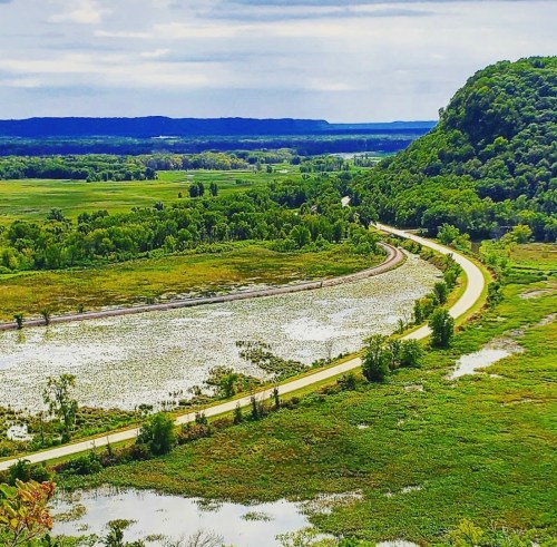 A winding road curves through lush green fields and wetlands, with hills in the background under a cloudy sky.