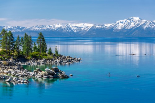 A serene lake with clear blue water, surrounded by snow-capped mountains and lush green trees, with kayakers in the distance.