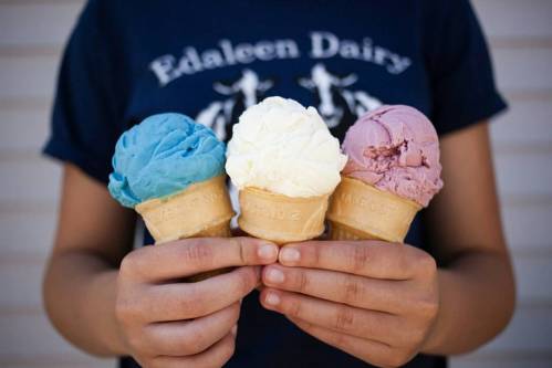 A person holds three ice cream cones: blue, white, and purple flavors, against a light background.