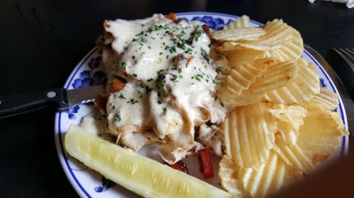 A plate of cheesy chicken casserole topped with herbs, served with potato chips and a pickle slice.