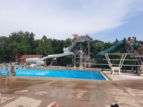 A sunny pool area with water slides, people swimming, and lounging on the deck surrounded by trees.