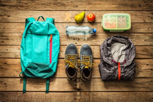 A teal backpack, shoes, a water bottle, fruit, and a jacket arranged on a wooden surface.