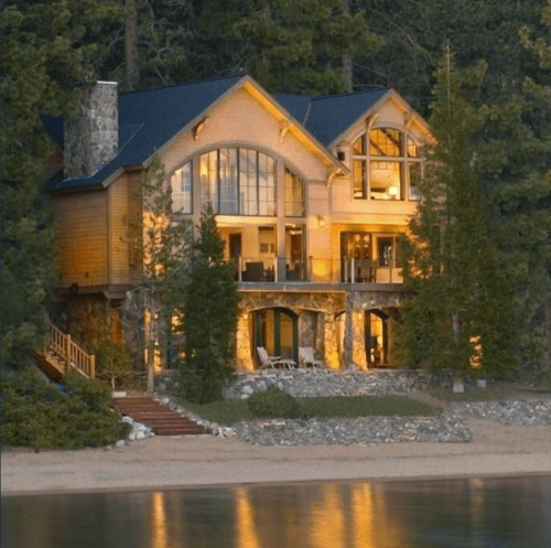 A large, modern lakeside house with large windows, surrounded by trees and a sandy beach. Warm lights illuminate the exterior.