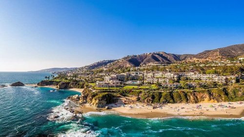 Aerial view of a coastal beach with clear blue water, sandy shore, and hillside homes under a bright blue sky.