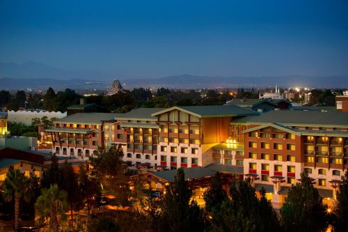 A hotel illuminated at dusk, surrounded by trees and mountains in the background, with city lights twinkling.