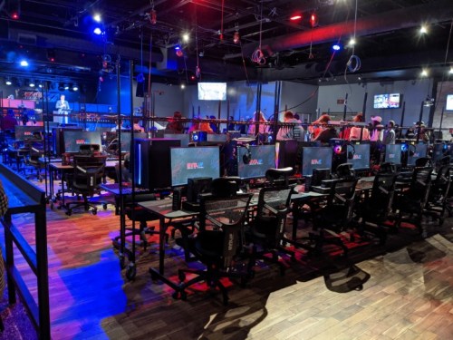 A spacious gaming lounge with rows of gaming stations, dim lighting, and screens displaying various games.