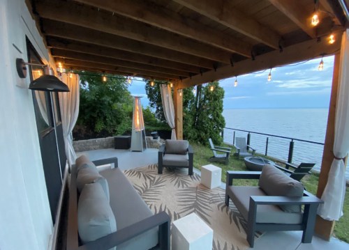 Cozy outdoor seating area with a view of the water, featuring a rug, chairs, and string lights under a wooden pergola.