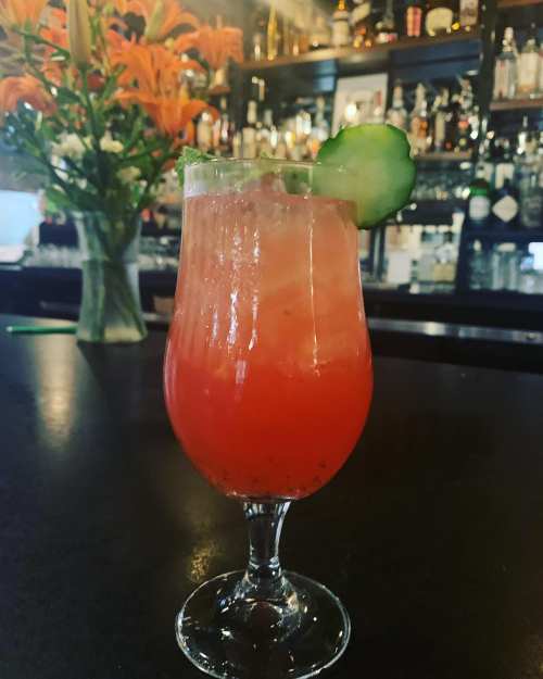 A refreshing cocktail in a tall glass, garnished with cucumber, set against a bar with bottles and flowers in the background.