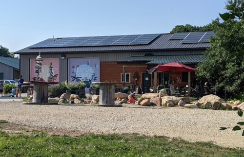 A rustic building with solar panels, outdoor seating, and a garden area, surrounded by gravel and rocks.