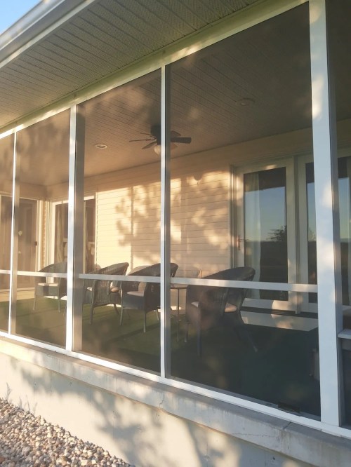 A screened porch with several chairs and a ceiling fan, surrounded by sunlight and greenery.