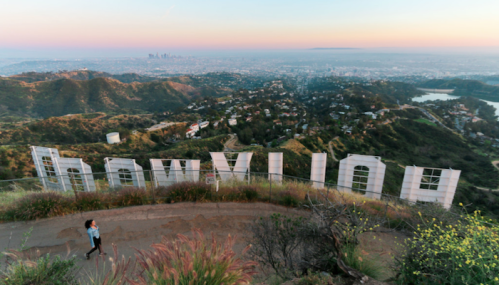 A person walks near the Hollywood sign, overlooking a cityscape at sunset with hills and greenery.