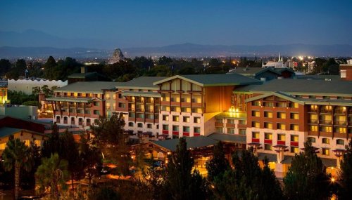 A hotel illuminated at night, surrounded by trees, with distant mountains and city lights in the background.