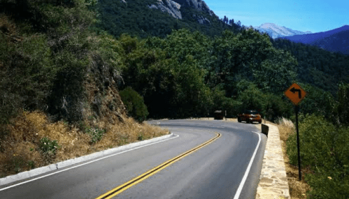 A winding road curves through a lush green landscape with mountains in the background and a car approaching a right turn.