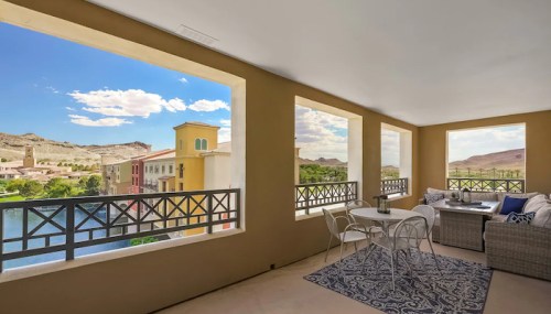 A spacious balcony with a table and chairs, offering a view of mountains and colorful buildings under a blue sky.