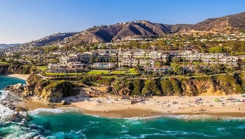 A scenic coastal view of a beach with waves, cliffs, and luxury homes on a hillside under a clear blue sky.