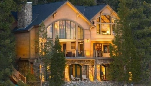 A modern wooden house with large windows, surrounded by trees, illuminated at dusk.