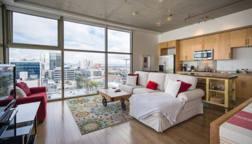 Modern apartment interior with a cozy living area, large windows, and city views. Light wood floors and stylish decor.