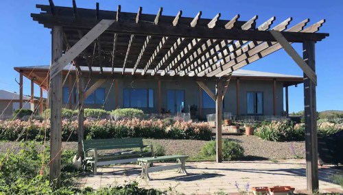 A wooden pergola with a bench in a garden, overlooking a modern house and landscaped greenery under a clear blue sky.