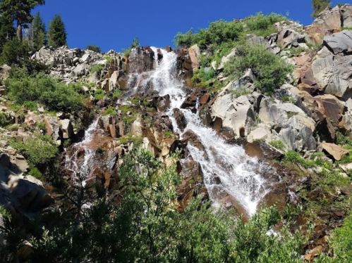 A cascading waterfall flows over rocky terrain, surrounded by lush greenery and a clear blue sky.