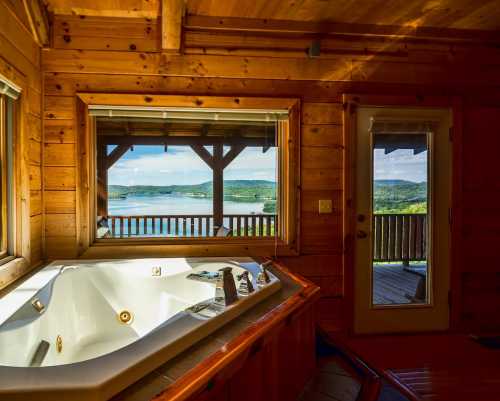 A cozy wooden cabin interior featuring a hot tub and a scenic view of a lake and hills through large windows.