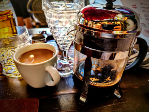 A French press with brewed coffee beside a small cup of coffee and a glass of water on a wooden table.
