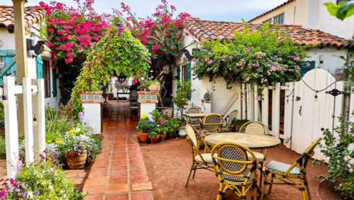 Charming courtyard with vibrant flowers, greenery, and outdoor seating in a sunny, inviting atmosphere.