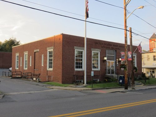 A brick building with large windows, labeled as a U.S. Post Office, located on a street with power lines and flags.