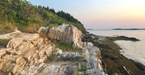 Rocky shoreline with colorful cliffs, lush greenery, and calm waters under a pastel sky at sunset.