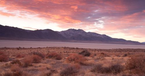 A desert landscape at sunset, featuring mountains in the background and colorful clouds in the sky.