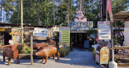 Entrance to a rustic barbecue restaurant with wooden animal sculptures and colorful signs in a wooded area.
